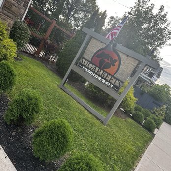 Sugarbeets - The image shows an outdoor sign for Sugarbeets Kitchen & Bar, positioned on a grassy area with small, rounded bushes in the foreground. The sign features a tree logo and the restaurant's name, with a wooden frame and a light fixture above it. Behind the sign, there are trees, a fence, and a sidewalk, with an American flag flying nearby. The setting appears to be in a suburban or small-town area with a partly cloudy sky overhead. - New American Near Me - Rome, New York