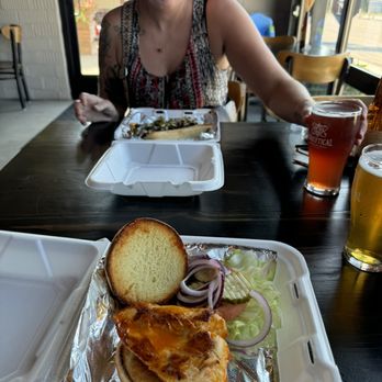 Analytical Brewing - A woman with light skin and dark hair, wearing a sleeveless patterned top, is seated at a table in a restaurant or cafe. She is reaching for a drink in a glass with a logo, while two takeout containers with food, including a sandwich with grilled chicken, lettuce, onions, and a slice of toasted bread, are on the table. The setting has large windows and wooden chairs, with sunlight coming in. - Beer Bar Near Me - Lexington, Illinois