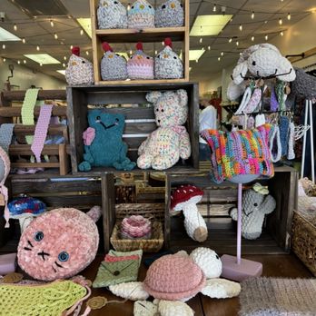Junk Head - Adorable knitted animals! - The image shows a variety of handmade knitted and crocheted stuffed animals and accessories arranged on wooden shelves and tables in a craft market setting. Items include chickens, frogs, bears, a cat, mushrooms, and various small accessories like bags and hats. The display features colorful and textured yarn creations, with some items on stands and others laid flat. The background includes ceiling lights and other market stalls, indicating an indoor craft fair environment. - Used, Vintage & Consignment Near Me - Mesa, Arizona