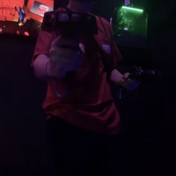 Krypton VR Lounge - Playing games! - A person wearing a virtual reality headset is taking a selfie in a dimly lit environment, holding a controller in one hand and a phone in the other. The background displays colorful screens with digital graphics, and the image includes the text 'CHICAGO JULY 02' and the timestamp '1:29 PM'. The scene suggests an entertainment or gaming setting, possibly at a VR lounge or arcade. - Virtual Reality Centers Near Me - Chicago, Illinois