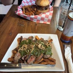 Chimichurri steak and fries and a rodeo burger - A burger with fries is served on red checkered paper, and a steak topped with onions and greens is on a white plate on a wooden table.