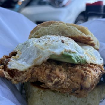 The West Egg - A breakfast sandwich featuring fried chicken, a fried egg, and avocado on a biscuit, with cars visible in the background. - Coffee & Tea Near Me - St. Augustine, Florida