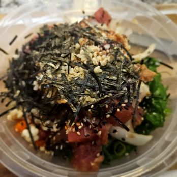 Poke Bros - a salad in a plastic container - A poke bowl in a transparent plastic container contains raw fish, shredded seaweed, sesame seeds, and chopped vegetables, representing a typical poke restaurant dish. The bowl is placed on a wooden surface, with the ingredients arranged in layers, highlighting the fresh and colorful presentation of the seafood and toppings. - Poke Near Me - Naperville, Illinois