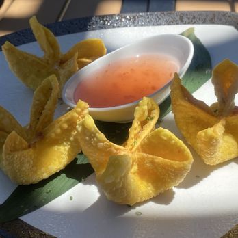 Hush - Crab Rangoon - Plate of fried wontons arranged around a small white dish of pink dipping sauce. The wontons are golden and crispy, placed on a white plate with a green leaf garnish underneath. The dish appears to be served outdoors, with sunlight casting shadows on the plate. - Sushi Bars Near Me - Keller, Texas