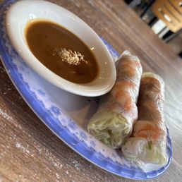 Spring Rolls - Two Vietnamese spring rolls filled with shrimp and vegetables are wrapped in translucent rice paper and placed on a blue and white patterned plate. A small bowl of dipping sauce, possibly hoisin or peanut sauce, is positioned next to the spring rolls. The plate is on a wooden table, with chairs visible in the background, indicating a restaurant setting.