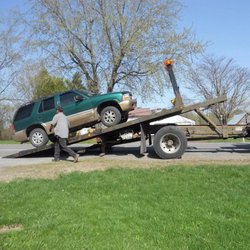 Gallagher's Towing & Auto - A green SUV is being loaded onto a flatbed tow truck by a person on a grassy roadside under a bright blue sky.