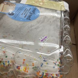 A cake with white frosting and multicolored sprinkles is inside a clear plastic container with a sealed lid. The container is placed in a cardboard box, and there are labels and stickers on the packaging. The cake appears to be pre-packaged for sale or delivery.