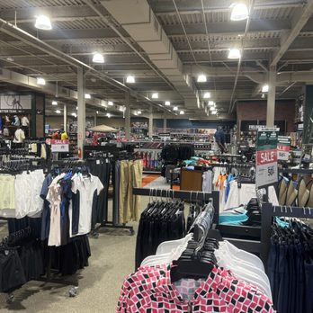 DICK'S Sporting Goods - The image shows clothing racks with various athletic and casual apparel inside a large retail store. Sale signs indicate discounts of 25% and 50%. The store has a high ceiling with exposed ductwork and bright overhead lighting. In the background, shelves and displays of sports equipment and accessories are visible, along with a few shoppers browsing the merchandise. - Sports Wear Near Me - Massapequa, New York