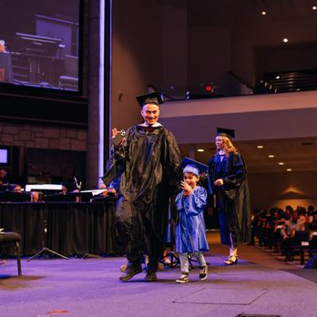 Amberton University - A working adult graduate celebrates on stage with a child during Amberton University's commencement ceremony as faculty and guests look on. - Colleges & Universities Near Me - Frisco, Texas Amberton University - A working adult graduate celebrates on stage with a child during Amberton University's commencement ceremony as faculty and guests look on. - Colleges & Universities Near Me - Frisco, Texas