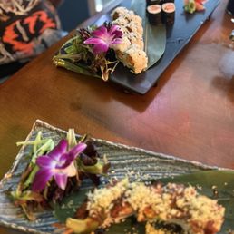 Two rectangular plates of sushi are displayed on a wooden table, each garnished with edible flowers and greens. The plate in the foreground has a partially eaten sushi roll with a crunchy topping, while the background plate features assorted sushi rolls, including maki and nigiri, with wasabi and pickled ginger. The setting appears to be in a Japanese restaurant or sushi bar.