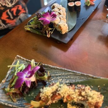 Sunflower Cafe - Two rectangular plates of sushi are displayed on a wooden table, each garnished with edible flowers and greens. The plate in the foreground has a partially eaten sushi roll with a crunchy topping, while the background plate features assorted sushi rolls, including maki and nigiri, with wasabi and pickled ginger. The setting appears to be in a Japanese restaurant or sushi bar. - Sushi Bars Near Me - Valrico, Florida