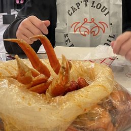 Snow Crab - A seafood boil with crab legs, sausages, and other seafood is placed on a table, covered with a plastic liner. A person wearing a bib with 'Cap't Loui' printed on it is reaching for the food, with their hands visible. The setting appears to be a restaurant with a casual dining atmosphere, including a red cup and utensils on the table.