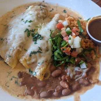 La Cueva Cafe - My yummy vegetarian enchiladas - A plate with vegetarian enchiladas covered in melted cheese and sauce, served alongside rice, beans, and a fresh salsa mixture with chopped vegetables and herbs. A small cup of dark sauce is also present on the plate, with some chopped herbs sprinkled on top. - Mexican Near Me - Taos, New Mexico