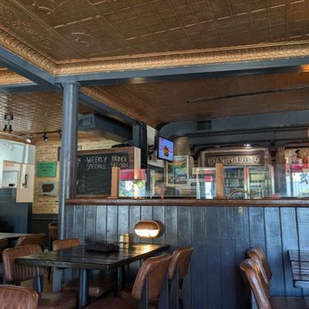 J Murphys on Main - Dining area - The image shows the interior of a restaurant with dark wood paneling on the walls and ceiling. Several tables with chairs are arranged throughout the space. A bar area with a refrigerator and bottles is visible in the background, along with a chalkboard menu and a small television. The lighting includes a hanging bulb and a small wall-mounted light fixture. - American Near Me - Cold Spring, New York