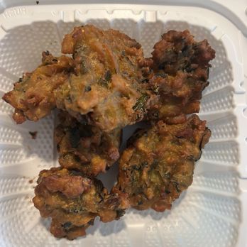 Monsoon Indian Kitchen - Veggie Korma - Four vegetable fritters, possibly vegetable pakoras, are arranged in a white plastic container with a textured surface. The fritters are golden-brown with visible bits of vegetables and herbs, indicating they are deep-fried and crispy. The container is set on a surface that appears to be a granite countertop. - Indian Near Me - Needham, Massachusetts