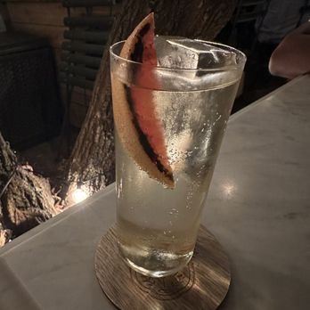 Bar Pompette - A transparent glass filled with a clear, bubbly drink, garnished with a lemon wedge, placed on a round wooden coaster on a bar counter. - Cocktail Bars Near Me - Toronto, Ontario