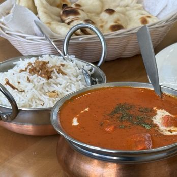 Indian hot breads - The whole shabang! - The image shows a meal with a bowl of white rice topped with fried onions, a bowl of red curry with herbs, and a basket of naan bread lined with paper, all placed on a wooden table. The curry appears to contain chunks of meat or vegetables, and a spoon is resting in the curry bowl. The naan bread is in a white basket with a handle, and a white plate is partially visible on the right side of the image. - Indian Near Me - Hamden, Connecticut