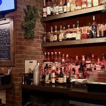 Wits End Saloon - Fully stocked bar - A bar shelf features multiple bottles of alcohol, including whiskey, flavored spirits, and liqueurs, arranged on three levels. The background includes a brick wall and a small decorative plant. The bar counter below holds additional bottles, cups, and bar tools, with a chalkboard menu visible on the left side and a television screen in the upper left corner. - Bars Near Me - Timonium, Maryland