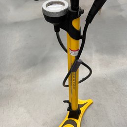 It's $10 cheaper BRAND NEW! - A yellow Serfas brand hand pump with a built-in pressure gauge and attached hose is positioned upright on a gray concrete floor. The pump features a black handle and a base designed for stability. A price sticker is visible on the pump, indicating it is for sale. The background is plain, with no additional objects or signage visible.