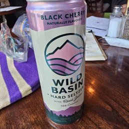 A tall, cylindrical can of Wild Basin Black Cherry flavored hard seltzer is positioned on a wooden restaurant table. The can features a mountain and landscape illustration in pink and black, with text indicating it is a naturally flavored, low-calorie beverage with 100 calories. The background includes menus, salt shakers, and other dining items, with patrons seated at tables in a casual dining setting.