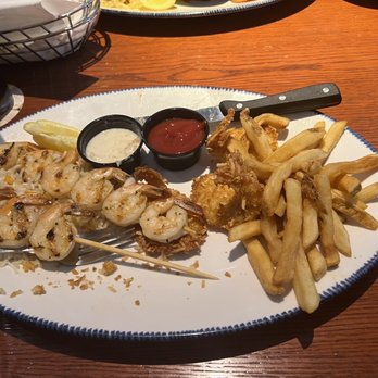 Red Lobster - Endless Shrimp with Parrot Isle Jumbo Coconut Shrimp and Grilled Shrimp Skewer - A seafood platter includes grilled shrimp on skewers, French fries, and two small containers of dipping sauces, one with tartar sauce and the other with ketchup. The dish is served on a white oval plate with a blue rim, placed on a wooden table. Additional food items and a glass of dark beverage are visible in the background. - Seafood Near Me - Albuquerque, New Mexico