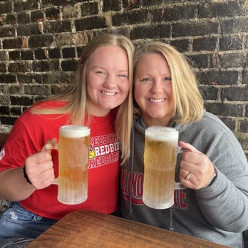 Brewe-Ha's - Go Redbirds! - Two women sit at a table in a bar, smiling and holding large mugs of beer, with a brick wall behind them. - Bars Near Me - Normal, Illinois