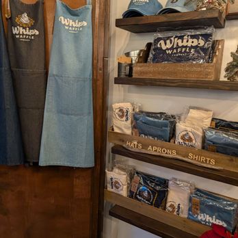 Whips Waffle - Aprons, hats, and other merchandise with the Whips Waffle logo are arranged on shelves and hanging on a wooden panel in a retail space. - Waffles Near Me - St. Augustine, Florida