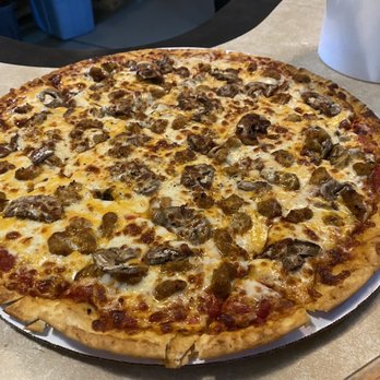 Cedar Shack - Dairyland pizza thin crust with sausage and mushroom. - A pizza with a golden-brown crust topped with melted cheese, sausage pieces, and sliced mushrooms. The pizza is round and appears freshly baked, sitting on a table surface with a paper pizza tray underneath. Part of a white container is visible in the background. - Bars Near Me - Adams, Wisconsin