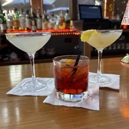 Three cocktails are arranged on a wooden bar counter, with two martinis—one with a lemon twist and one with a lemon wedge—and a Negroni with an orange slice and a black straw. The background shows a bar with bottles, a window, and some decorative elements, creating a lively bar scene.