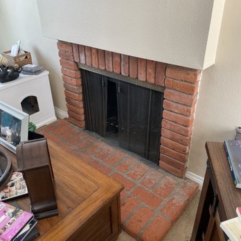 OC Fireplace Pro's - Before - A brick fireplace with a white mantel is located in a living room, featuring a brick hearth extending onto the carpeted floor. The fireplace opening is covered with a black mesh screen. Nearby furniture includes a wooden table, a white cabinet, and various decorative items and books. The wall above the mantel is painted in a light color, and a framed picture is partially visible at the top of the image. - Fireplace Services Near Me
