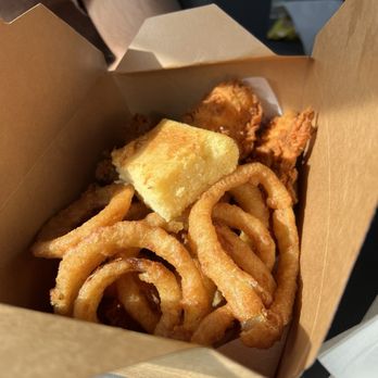 Basha's Chicken - A brown paper takeout box contains fried onion rings, fried chicken pieces, and a piece of cornbread. The onion rings are golden and crispy, while the chicken pieces are breaded and fried. The cornbread is a small, square piece with a golden crust. The background shows part of a person's arm and a gray surface, indicating a casual dining or takeout setting. - Chicken Shop Near Me - Warren, Michigan