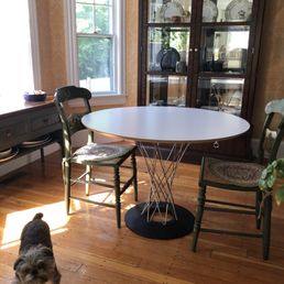 My dog can vouch for the fact that this is a great table! - The image shows a dining room with a round white table at the center, surrounded by two green chairs with floral cushions. A dark wood china cabinet with glass doors displays plates and decorative items. There is a window with white trim allowing natural light into the room, and a small dog is standing on the wooden floor near the table. The room has beige wallpaper and hardwood flooring.