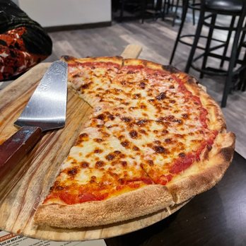 Pizzeria Bruno - Cheese pizza with a golden, slightly charred crust on a wooden board, served in a restaurant with black chairs and tables in the background. - Italian Near Me - Sutton, Massachusetts
