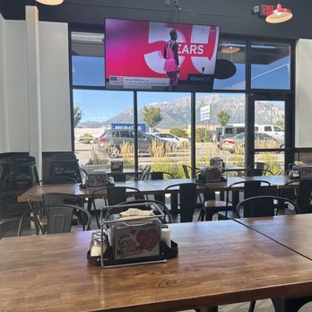 Mountain Mike's Pizza - The image shows the interior of a restaurant with multiple wooden tables and black chairs arranged for dining. A large flat-screen TV is mounted on the window wall, displaying a sports event with a person in a pink outfit. Outside the window, parked cars and mountains are visible. The restaurant has modern lighting fixtures, including red pendant lights, and a condiment holder on the table. The setting appears bright with natural light coming through the windows. - Pizza Near Me - Orem, Utah