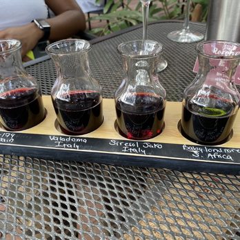 Bin 39 - International flight - Four small glass wine bottles with dark red and green liquids are arranged on a wooden tray with handwritten labels indicating their origins from Spain, Italy, Italy, and South Africa. - Wine Bars Near Me - St. Augustine, Florida