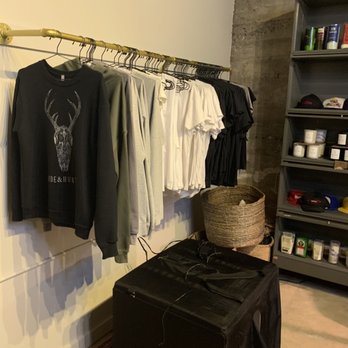 Hide & Hunter - Hoodies and t-shirts in black, white, and gray are displayed on a gold clothing rack in a retail or storage area, with a black speaker and storage shelves nearby. - Leather Goods Near Me - Birmingham, Alabama
