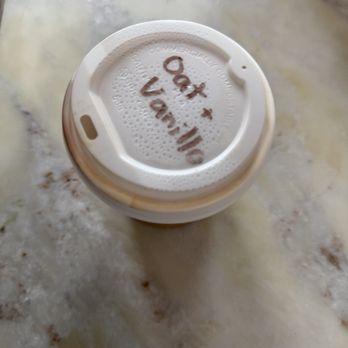 Creative Grounds DC - A disposable coffee cup with a plastic lid is positioned on a marble surface. The lid has handwritten notes in black ink, including the words 'Oat' and 'Vani Mo,' along with some symbols. The marble surface has a light color with subtle veining and some small spots or stains. - Art Classes Near Me - Washington, DC, District of Columbia