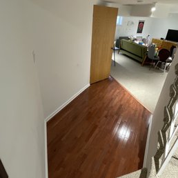 Final result - The photo captures a view from the top of a staircase overlooking a living room area. The staircase has a carpeted edge, and the floor below is made of polished wood. A wooden door is partially open, revealing a room with a green sofa, a dining table with chairs, and various household items. The room has white walls, a small window, and a television on a stand. The space appears to be part of a residential interior.