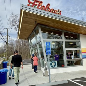 Flobee's Roadside Stand - Flobee's - The image shows a group of people standing outside Flobe's ice cream shop, which features a modern architectural style with large glass windows and a wooden ceiling overhang. The shop has a sign indicating reserved parking and accessibility features. The parking lot is visible with marked spaces, and there are trees and power lines in the background. The sky is partly cloudy. - Fast Food Near Me - Ridgefield, Connecticut