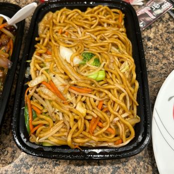 My Kitchen - Vegetable lo main - A black plastic takeout container contains vegetable lo mein with cooked noodles mixed with vegetables such as carrots, broccoli, cabbage, and bean sprouts. The container is placed on a speckled brown countertop, with small cups of red sauce and other food items visible in the background. The dish appears to be a Chinese-style noodle entrée served in a fast-casual setting. - Chinese Near Me - Gilbert, Arizona