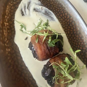 Pilon Miami - Appetizer apricots. Super tasty. - Bacon-wrapped dates topped with microgreens are presented on a textured, dark-colored plate with a creamy white sauce. - Latin American Near Me - Miami, Florida