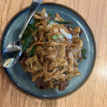 Chef Liu's Kitchen - Beef Chow Fun - A serving of beef chow fun is presented on a blue ceramic plate, containing stir-fried flat rice noodles, beef slices, and green vegetables such as scallions. A metal spoon rests on the plate, and the dish is placed on a wooden table. The noodles appear glossy and well-cooked, with visible pieces of beef and vegetables mixed throughout. - Chinese Near Me - Aurora, Colorado