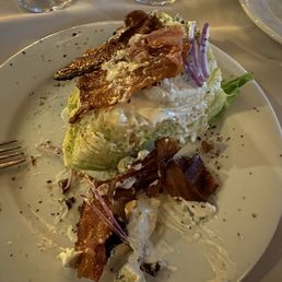 A partially eaten salad featuring lettuce, bacon strips, grilled fish, and sliced red onions on a white plate. The dish appears to have been served with dressing, with some food remnants and scattered pepper or seasoning. The plate is set on a table with wine glasses and additional plates visible in the background.