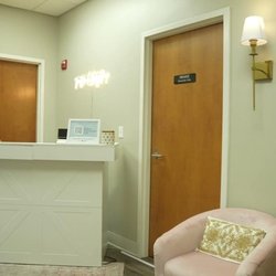 A brightly lit, modern reception area with a white counter, a pink armchair, and two wooden doors...