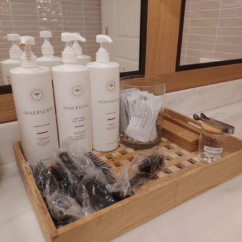 Willowbrook Spa - Hair care/toiletries - A wooden tray on a bathroom countertop holds three large bottles of hair care products labeled 'InnerSense,' a clear container with wrapped items, a small jar labeled 'Dental Tabs,' and some black hair accessories in plastic. The background features a mirror, tiled wall, and a white door, indicating a bathroom setting. - Estheticians Near Me - Canandaigua, New York