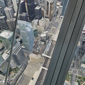 CN Tower - From level 147 - The image captures a city skyline from a high vantage point, likely from an observation deck of a tall building. It shows numerous skyscrapers of varying heights and architectural styles, with streets and railway tracks visible below. The photo includes glass and metal structural elements of the observation area, indicating it was taken from a modern high-rise building. - Landmarks & Historical Buildings Near Me - Toronto, Ontario