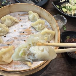 Dumplings are arranged in a bamboo steamer, with one being held by chopsticks, surrounded by dipping sauces and empty plates.