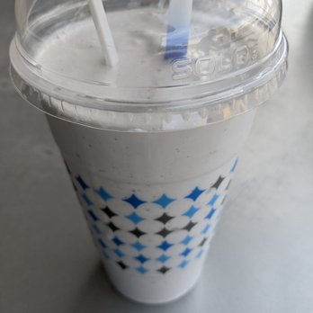 Frost Gelato - Milk Shake, (Grande size), $6.75 prior to tax - A milkshake in a tall, branded cup with a clear plastic lid and two straws, sitting on a light-colored surface. The cup features a geometric pattern in black, white, and blue. The milkshake appears to be cookies and cream flavor, with visible cookie bits mixed into the creamy beverage. - Desserts Near Me - Albuquerque, New Mexico