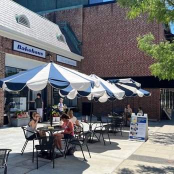 Bakehaus - Outside - Outdoor patio with tables, chairs, and large striped umbrellas outside Bakehaus bakery, with customers dining and a sidewalk sign. - Desserts Near Me - Belmont, Massachusetts