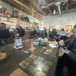The image shows a cafe or bar interior with multiple patrons seated at tables and the bar. Some customers are engaged in a card game on a table, with cards and game boxes visible. The space features an industrial ceiling with exposed ductwork, hanging lights, and artwork on the walls. The bar area has a menu and potted plants, and patrons are using their phones or chatting. The environment appears lively and social.